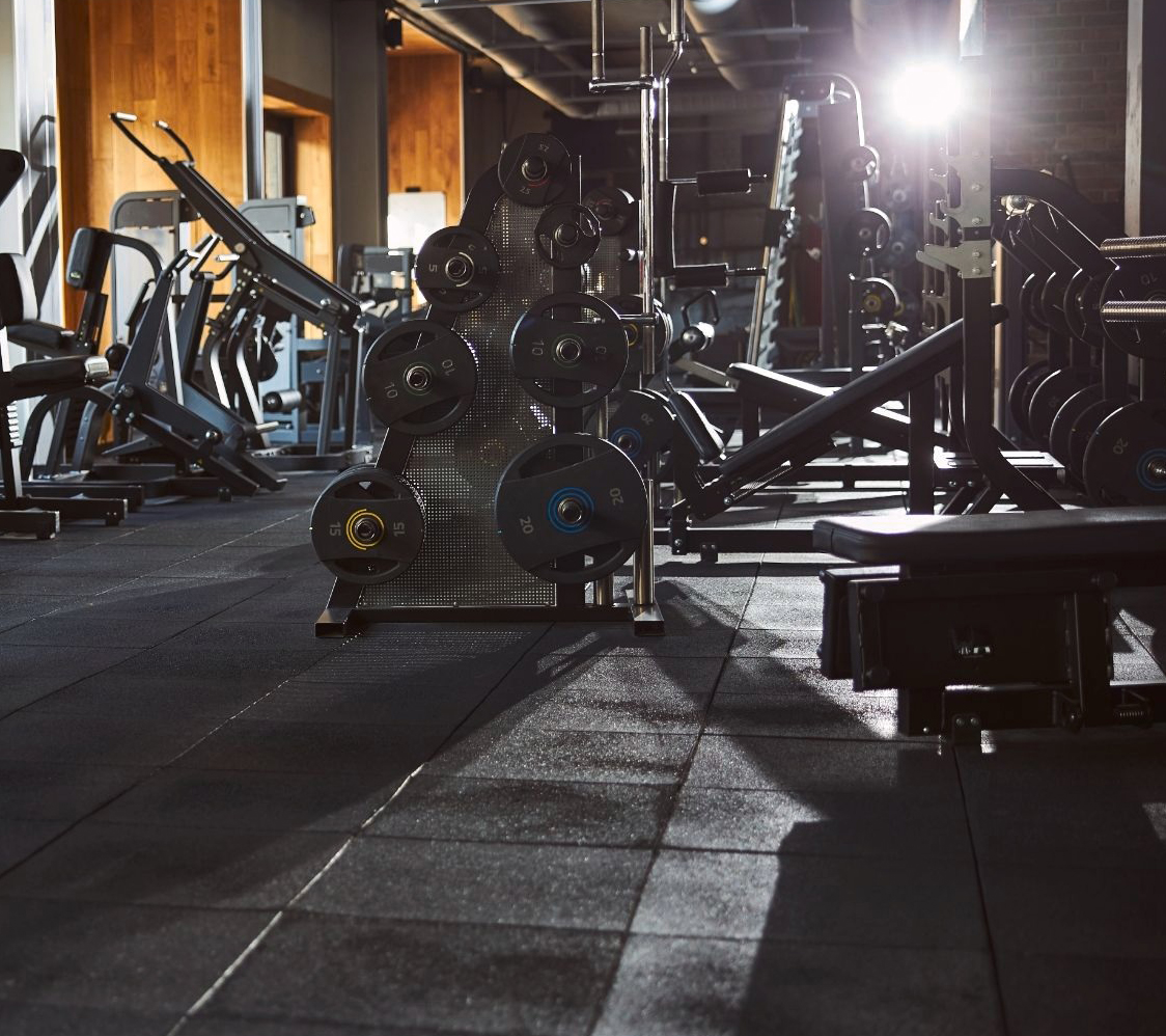 A gym with various weight machines, benches, and a rack holding multiple weight plates. The room is empty, with sunlight streaming in, casting shadows on the black rubber floor.