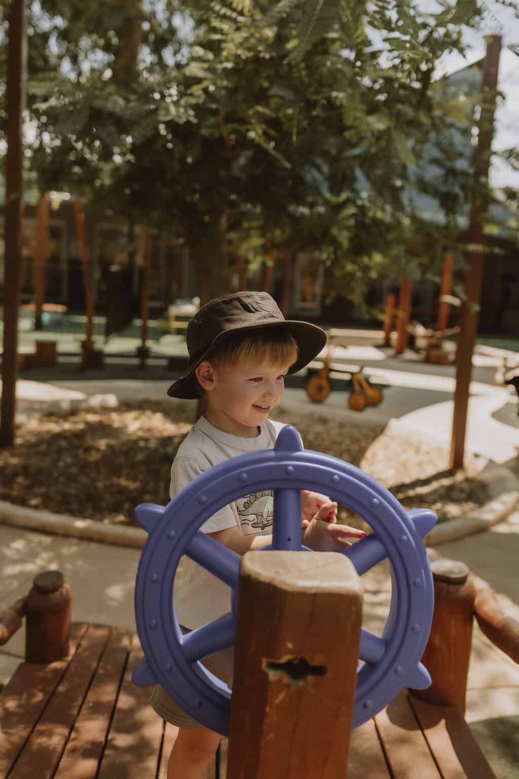 A boy in a bucket hat steering the blue ship's wheel in the Cuddles Kelmscott playground
