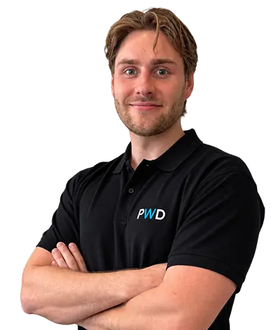 Luke, with short swept-back hair and light stubble, smiles at the camera with arms crossed, wearing a black PWD polo shirt against a plain white background.